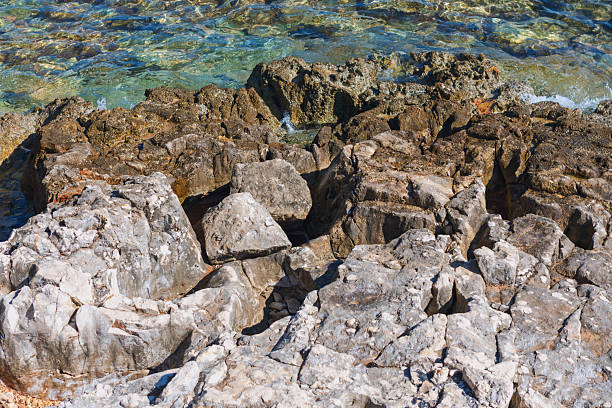 Rocky coastline in Croatia Mediterranean region, featuring large boulders at shore meeting turquoise water. Scene showcasing interplay of sunlit, rugged rock textures and the shallow, transparent sea