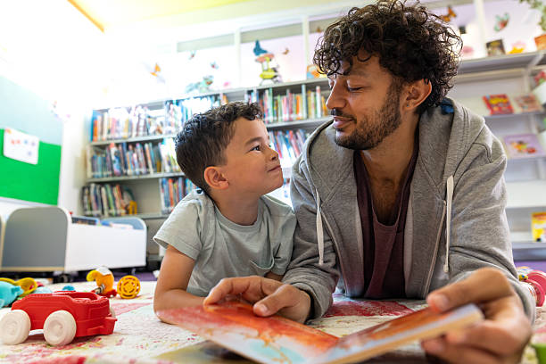 Medium shot of a father and son reading a story together while lying on the floor in a community library in Newcastle-upon-Tyne, North East England. They are both looking at each other affectionately. There are bookshelves and toys visible in the background, out of focus.Videos similar to this scenario available.