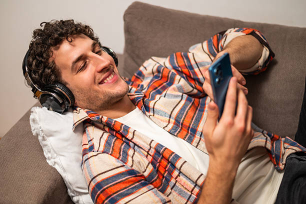 istock Smiling young man using headphones and phone while relaxing on sofa at home 2256418209