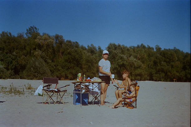 A mother is standing beside her son and daughter who are sitting on folding chairs on a sandy river beach during a family day trip in Serbia, photographed in daytime on 35mm analog film with an analog camera.