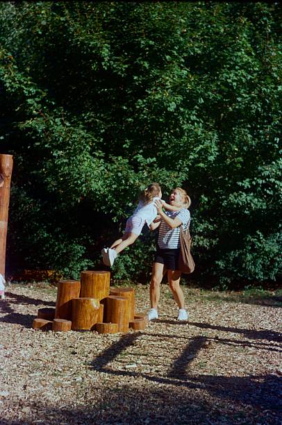 A mother is lifting her young daughter while playing outdoors in a park setting surrounded by trees, photographed during daytime on 35mm analog film with an analog camera.
