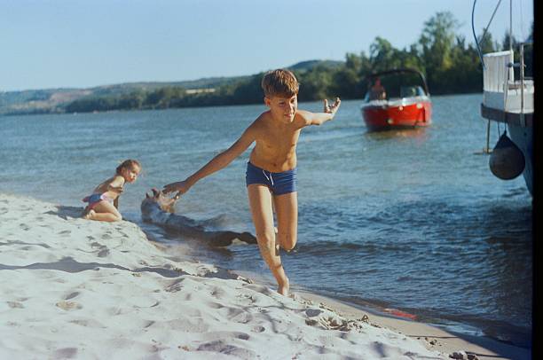 A brother and sister are playing in the sand by a river while standing near their family boat during a summer day trip in Serbia, photographed in daytime on 35mm analog film with an analog camera.
