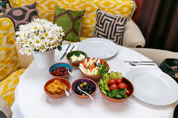 A beautifully arranged Mediterranean style breakfast served on a white tablecloth, featuring assorted cheeses, fresh tomatoes, cucumbers, olives, jams and butter presented in small bowls, complemented by a vase of white daisies and set in a cozy, stylish restaurant seating area, highlighting healthy eating, freshness and elegant morning dining atmosphere