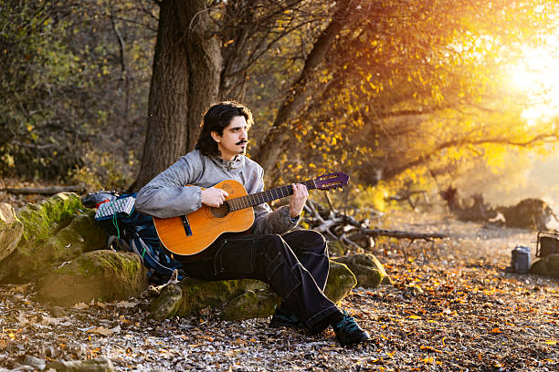 A young man with a handlebar mustache sits on mossy rocks in an autumn forest, playing an acoustic guitar during a golden sunset, with hiking gear nearby