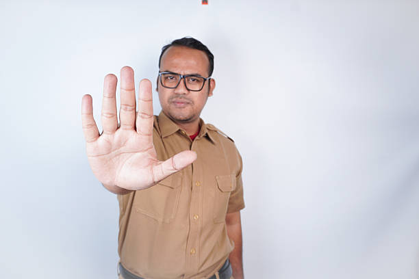 African american security officer raising a finger in front of the camera, showing a restriction symbol against blue background. Man working as a patrol agent presents a stop sign in studio.