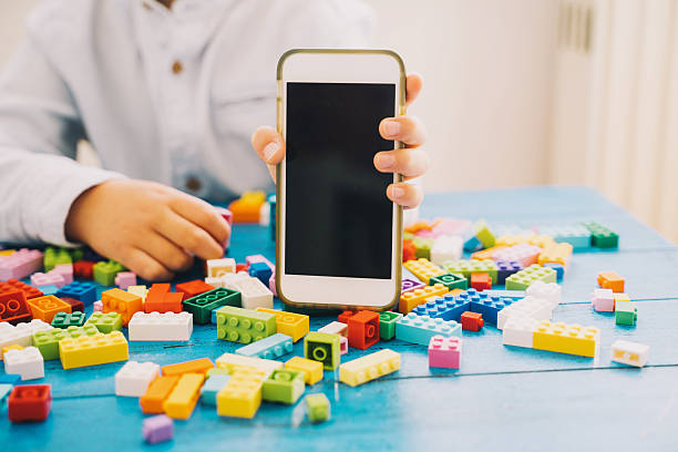 Mockup image of child hand holding and showing mobile phone with blank white screen with colorful toy bricks or plastic blocks. Kids leisure and education at home or class, learning and development.