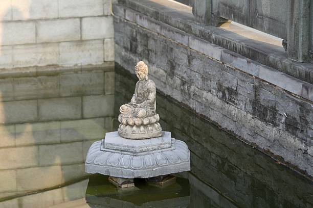 This peaceful image captures a weathered stone statue of a seated Buddha meditating on a lotus pedestal in a shallow reflective pond, surrounded by aged stone balustrades in a traditional temple setting. The soft natural light and subtle water ripples evoke tranquility, spirituality, and cultural heritage, ideal for wellness retreats, meditation apps, historical illustrations, or inspirational visuals in editorial, advertising, or spiritual wellness campaigns.