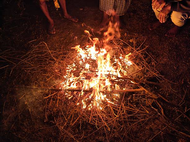 People warming their hands and feet over a glowing campfire. Bright orange flames flicker amidst charred wood and grey ash. This is a traditional way to keep the body warm during cold weather.