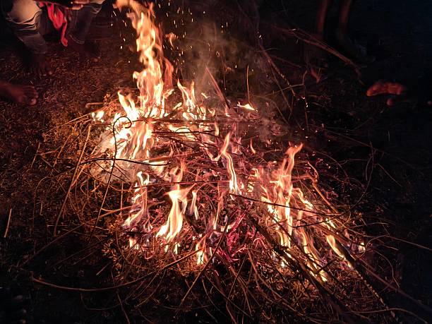 People warming their hands and feet over a glowing campfire. Bright orange flames flicker amidst charred wood and grey ash. This is a traditional way to keep the body warm during cold weather.