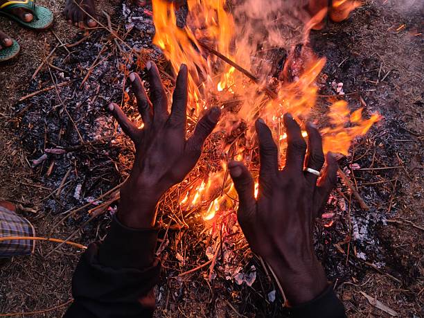 People warming their hands and feet over a glowing campfire. Bright orange flames flicker amidst charred wood and grey ash. This is a traditional way to keep the body warm during cold weather.