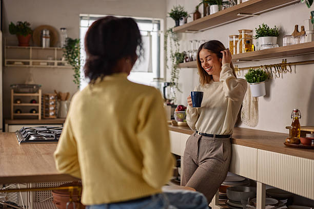 Friends chat in a cozy kitchen, warm lighting, plants, and coffee mug in hand