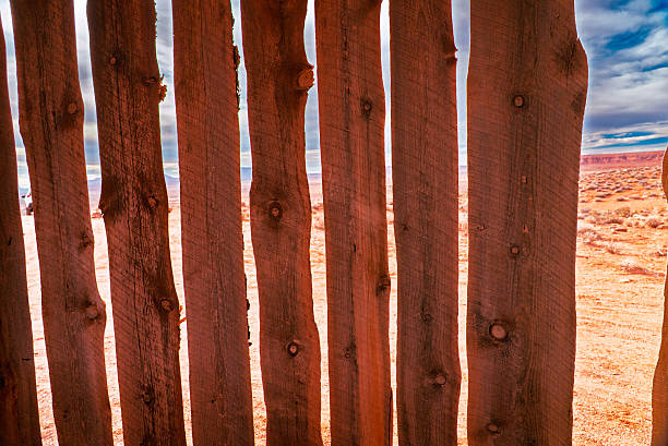 A close-up view of vertical, weathered wooden fence posts stands in the foreground, their rough textures, knots, and natural imperfections highlighted by warm desert light. Through the narrow gaps between the boards, an expansive red desert landscape is visible, stretching toward distant mesas beneath a partly cloudy sky. The image emphasizes contrast between manmade boundaries and the open, arid environment beyond, evoking themes of isolation, rural life, land division, and the interaction between human structures and the Southwestern desert.
