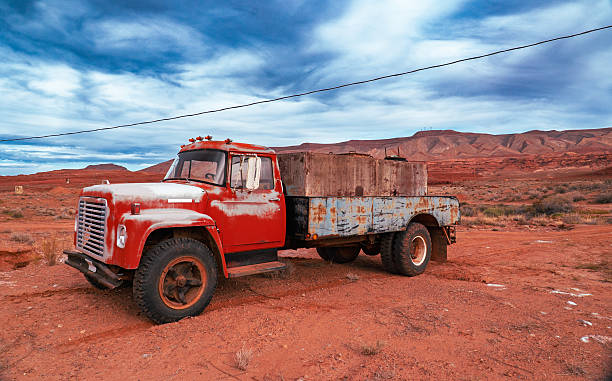 A weathered vintage flatbed truck with a faded red cab and rusted steel bed sits abandoned on red desert soil beneath a dramatic sky filled with swirling clouds. The truck shows heavy signs of age, corrosion, and long-term exposure to harsh desert conditions. Surrounding the vehicle are sparse desert shrubs, dry terrain, and distant layered red rock mesas typical of the American Southwest. The image conveys themes of industrial decay, abandonment, rural history, and the passage of time within an arid, remote landscape.