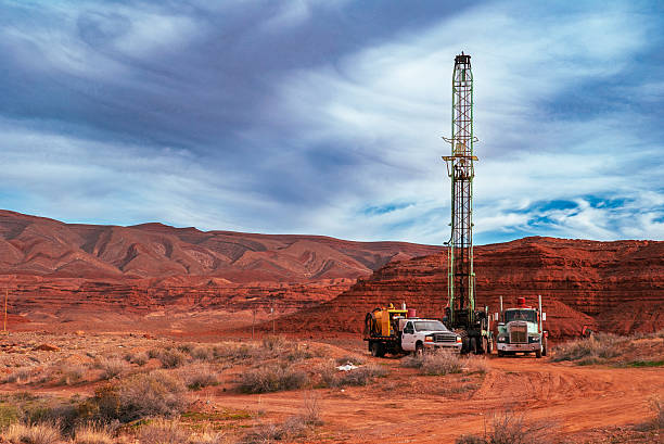 A vertical oil drilling rig stands at an active exploration site in a red rock desert basin, flanked by service trucks and drilling support equipment. Layered sandstone mesas and eroded canyon formations dominate the background, while dramatic cloud patterns sweep across the sky. The image illustrates industrial energy development set within a rugged, arid Southwestern landscape, emphasizing the scale of infrastructure against exposed geology.