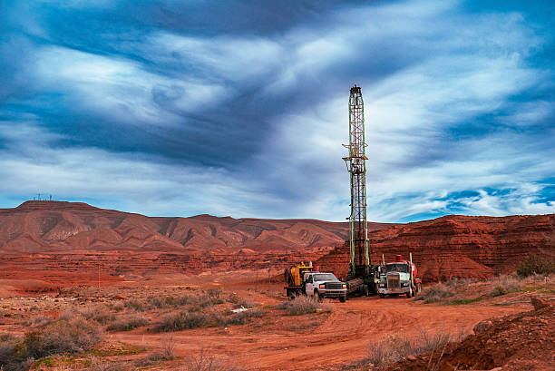An active oil drilling rig rises vertically from a red dirt access road in a remote desert basin, accompanied by service trucks and support equipment. Expansive red rock mesas and layered sedimentary formations stretch across the background, while dynamic, swirling cloud patterns dominate the sky overhead. The image captures the intersection of industrial energy exploration and rugged Southwestern geology, highlighting scale, isolation, and land use in an arid environment.