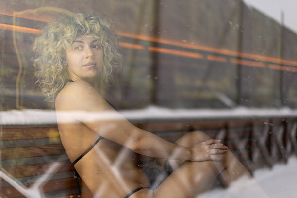 Beautiful woman wearing a bikini is sitting in a wooden sauna, relaxing, and looking through the window on snowy winter wonderland.