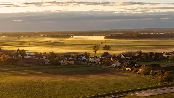Aerial view of a small village surrounded by expansive agricultural fields covered in morning mist. The scene captures the tranquility of the countryside as the mist lingers over the landscape. Houses and fields are visible under a soft morning light.