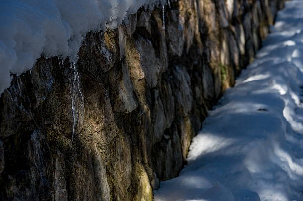 Snow covers the edge of a building while icicles hang down. The structure shows bricks and concrete. The sky appears blue and clear on a sunny winter day.
