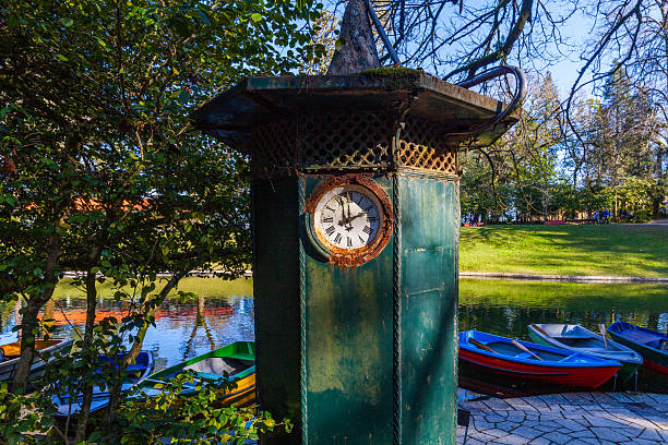 A weathered, green-painted clock tower stands by a tranquil lake, its rusted face showing the time, with colorful rowboats moored nearby and lush trees framing the serene park scene