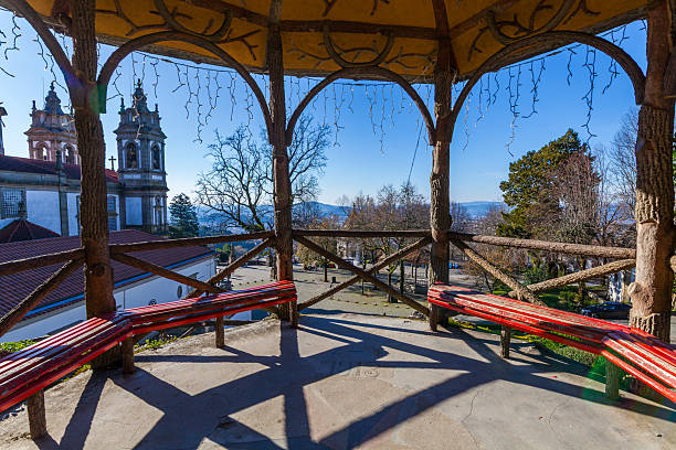 Viewed from inside a rustic, tree-trunk gazebo with red benches, a sunlit plaza and a historic church with twin towers are visible against a clear blue sky, casting long shadows on the ground