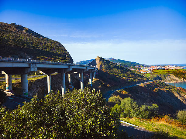 Modern road bridge crossing green coastal hills in Castro Urdiales, northern Spain. Scenic landscape with winding road, distant town, clear blue sky and natural vegetation. Infrastructure integrated into rugged coastal terrain. No people.