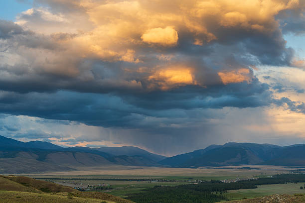 Wide view of an Altai valley with open steppe and distant mountain ridges under dramatic storm clouds, with visible rain shaft and warm sunset light breaking through. Large copy space in the sky makes it ideal for weather, climate, and adventure travel themes.