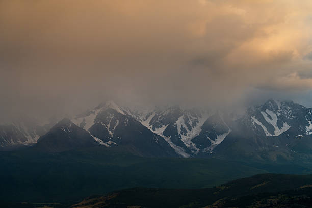 Epic panoramic view of the North-Chuya Range’s snow-covered peaks shrouded in mist and golden-hour clouds in Altai Republic, Russia, with dark green taiga foothills below creating moody atmospheric depth and copy space in the vibrant sky. Perfect for dramatic landscape, adventure, and wilderness photography.