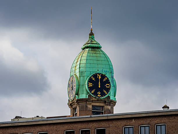 Copper dome of a historic clock tower with four clock faces, showcasing time and a directional wind vane against cloudy sky