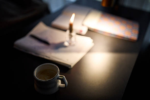 Cup of coffee placed on a table with a lit candle and an open notebook in dim lighting, creating a cozy and intimate atmosphere. The image represents relaxation, focus, writing, creativity, and a warm, moody workspace. Concept of home office, evening routine, coffee break, and inspiration.