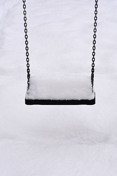 An empty black playground swing hangs from metal chains covered in a layer of white snow during a cold winter day