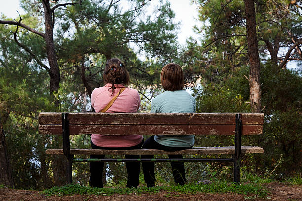 Caucasian young adult man sitting on bench with Caucasian young adult woman, kissing pet dog, while enjoying time together in forest park during autumn, sunlight filtering through trees