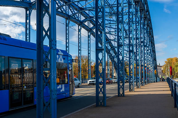 Krakow, Poland – October 25, 2025: A blue steel arch bridge spans the Vistula River as a city tram and cars move across it. The historic Grunwaldzki Bridge structure highlights everyday urban transport and engineering in Krakow.