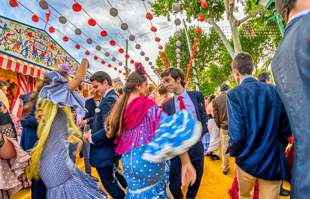 At the Seville April Fair, individuals in traditional attire perform the sevillanas dance near a decorated booth under paper lanterns during daylight hours.