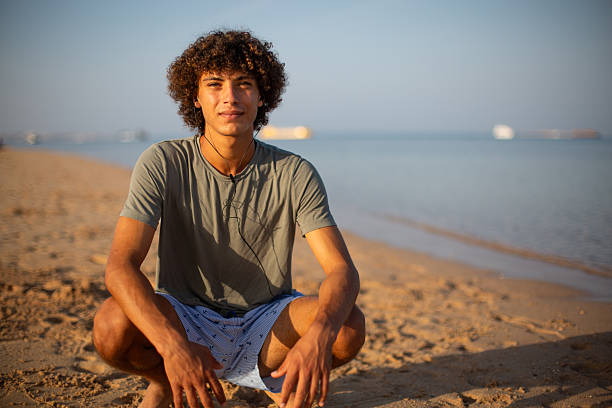 Young man smiling at beach while crouching on sand with ocean in background