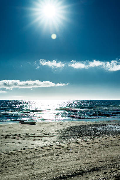 A brilliant sunburst radiates through a clear blue sky above a calm ocean, casting shimmering reflections across gentle waves and wet sand. An abandoned boat rests quietly along the shoreline, emphasizing solitude and stillness. Soft clouds stretch across the horizon while lens flare and high-contrast light create a cinematic, contemplative coastal scene ideal for wellness, travel, and reflective storytelling.