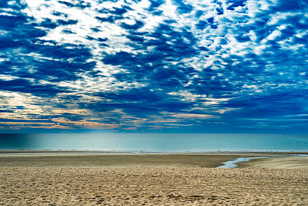 An expansive sandy beach stretches toward a calm ocean beneath a dramatic blue sky filled with dense, textured cloud formations. Shallow tidal water creates subtle leading lines across the sand, guiding the viewer’s eye toward the quiet horizon. The scene emphasizes scale, atmosphere, and natural contrast between land, sea, and sky, conveying a tranquil yet powerful coastal environment with no human presence.