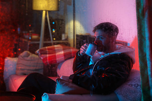 Young Man Drinking Hot Coffee at Home on Rainy Night with Neon Light Reflection on Window, Men's Lifestyle