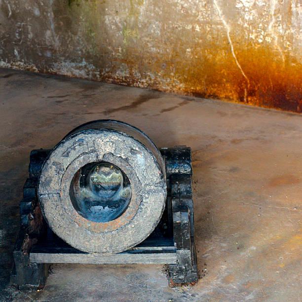 An aged cannon with a rugged stone wheel sits on a simple wooden platform in a dim, damp room with rusty walls, conveying history, abandonment, and weathered textures.
