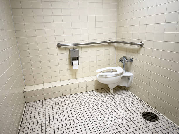 Wide angle view of an accessible public restroom featuring a wall mounted toilet, stainless steel grab bars, toilet paper dispenser and small trash bin against beige tiled walls and mosaic tile floor, captured in bright even lighting.​The clean but utilitarian design highlights hygiene, disability access, sanitation standards and public facility infrastructure suitable for editorial, safety and facility management themes.