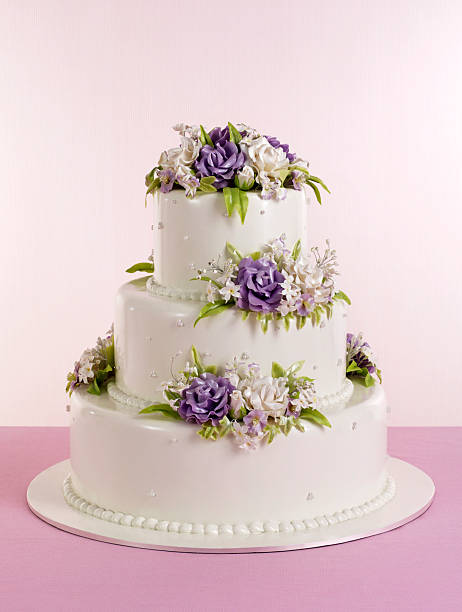 Front view of an elegant three tier wedding cake covered in smooth white fondant and adorned with clusters of purple and white blossoms on each level, displayed on a round cake board against a soft pink backdrop for a romantic look.​The clean styling, pastel colors and classic design make this image ideal for concepts related to weddings, bridal magazines, bakery promotions, event planning and celebration invitations.