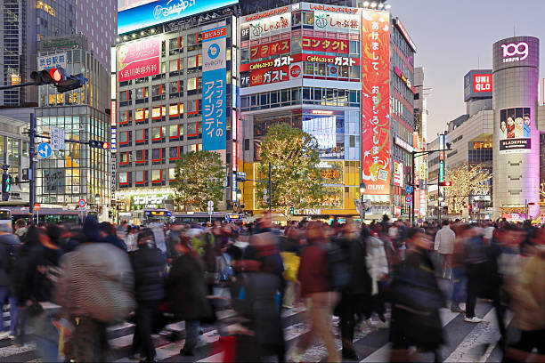 Shibuya Crossing at dusk in Tokyo, Japan. Large crowds of pedestrians move through the famous scramble intersection, surrounded by illuminated buildings, billboards, and city lights.