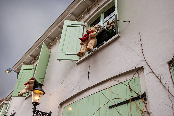 Three mint shuttered windows display teddy bears peeking and floating in balloon baskets, adding playful charm to a festive white facade with string lights and vines.