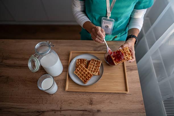 Healthcare worker in scrubs and id badge spreads strawberry jam on a golden waffle beside pitcher and glass of milk on a wooden table, conveying care, nutrition, and comfort