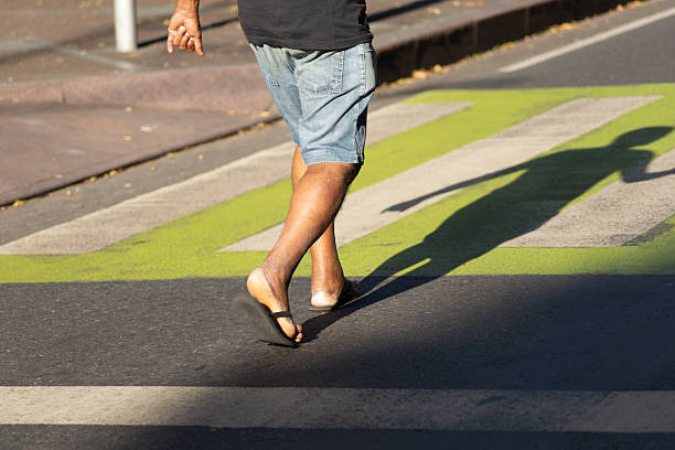 Below view, detail of the legs of a person wearing white pants and brown leather sandals, walking on asphalt with a green stripe.
