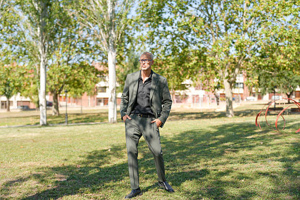 A man in a suit stands in a park. The park is filled with trees and has a playground