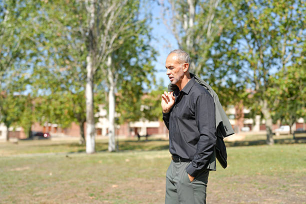 A man in a black shirt and gray pants is standing in a park. He is wearing a jacket and holding a cell phone. The scene is peaceful and calm, with trees in the background and a grassy field