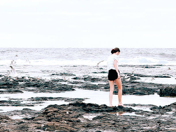 A young woman walks barefoot across rocky tidal pools as seabirds gather along the shoreline under an overcast coastal sky.