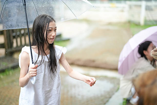 A girl is holding an umbrella and smiling. She is standing in the rain. There are other people in the background, some of them holding umbrellas as well