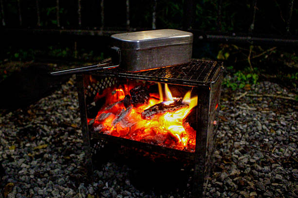 A close-up view of outdoor cooking with a traditional mess kit over a crackling bonfire. The bright orange flames and glowing embers create a warm atmosphere for a camping dinner. Ideal for outdoor lifestyle, bushcraft, and autumn or winter camping themes.