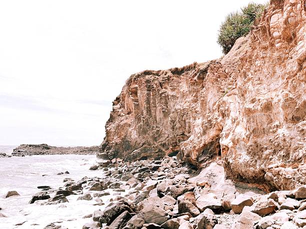 A rugged coastal cliff meets a rocky shoreline as waves wash over stone formations beneath a pale sky.